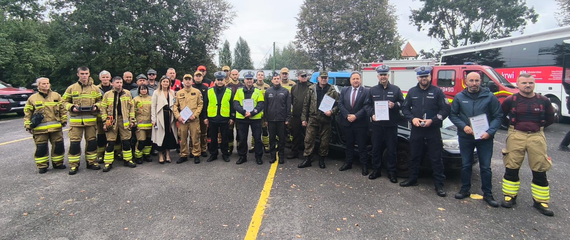 Na zdjęciu widoczni uczestnicy "Powiatowego Dnia Bez Ofiar Śmiertelnych na Drogach" w tym strażacy z Komendy Powiatowej Państwowej Straży Pożarnej w Głubczycach, Ochotniczej Straży Pożarnej w Dzierżysławiu, funkcjonariusze Wydziału Ruchu Drogowego Komendy Wojewódzkiej Policji w Opolu i Komendy Powiatowej Policji w Głubczycach, a także Sekretarz Wojewódzkiej Rady Bezpieczeństwa Ruchu Drogowego z Wojewódzkiego Ośrodka Ruchu Drogowego w Opolu.