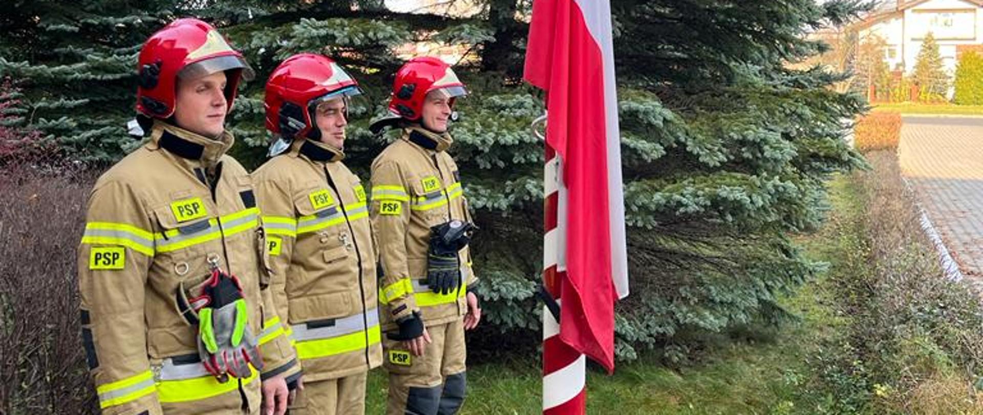 na zdjęciu widzimy trzech strażaków w ubraniach bojowych z hełmami na głowach stojących na baczność przed masztem ze sztandarem w tle drzewa