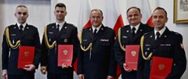 Pięciu strażaków w mundurach wyjściowych ze sznurem stoi obok siebie mężczyzna w środku jest bez teczki za nimi ustawione są trzy flagi Polski wisi obraz oraz znajdują się drzwi. 