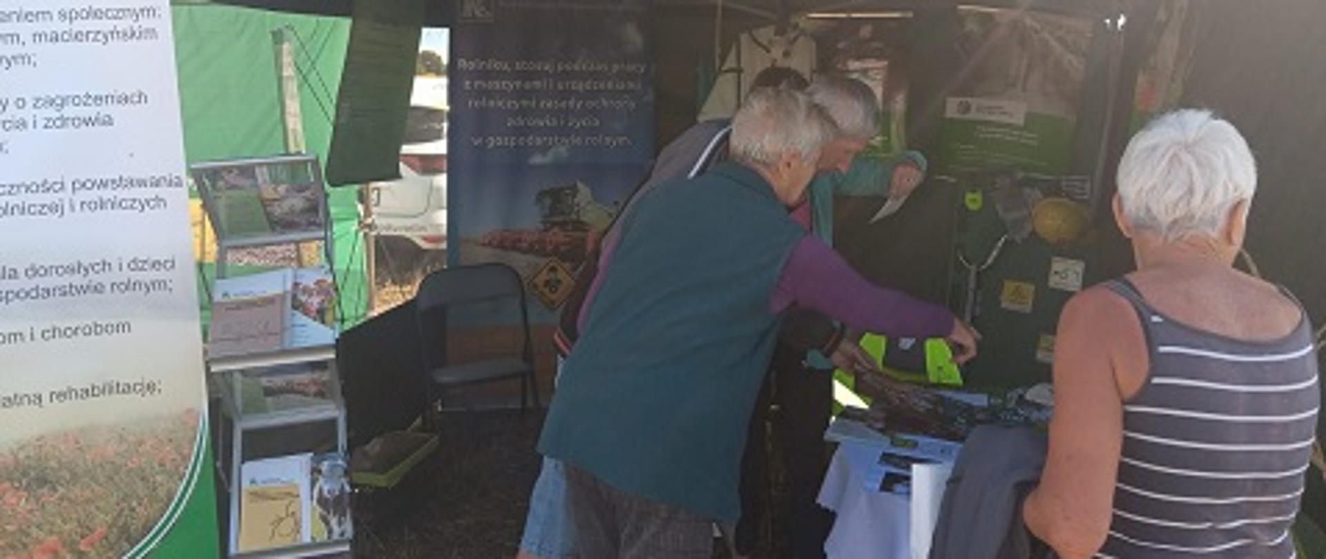 Przy zielonym namiocie KRUS stoją kobieta i dwóch mężczyzn, którzy oglądają wyłożone ulotki i broszury.