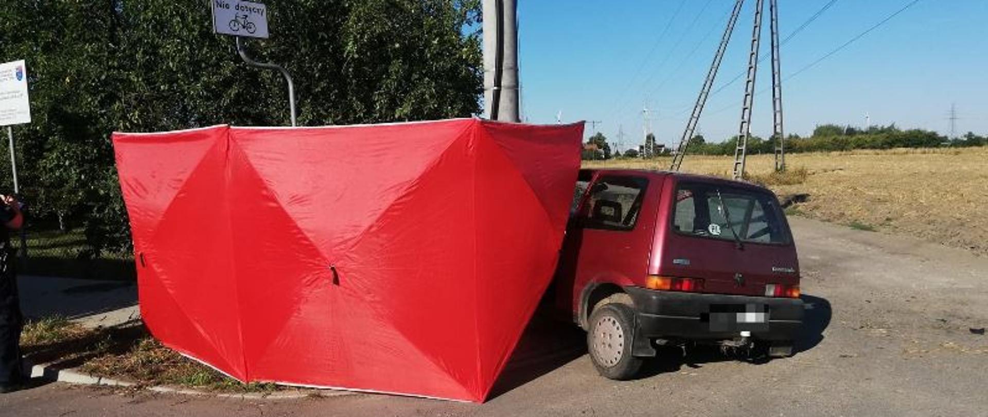 Część samochodu, który uderzył w słup widoczna poza czerwonym parawanem ratowniczym służb ratunkowych. Pojazd znajduje się na skrzyżowaniu drogi powiatowej z drogą gruntową. W oddali widoczne pole uprawne oraz drzewa owocowe rosnące na terenie działek rekreacyjnych w pobliżu słupa w który uderzył pojazd. Słoneczna pogoda i bezchmurne niebo. 