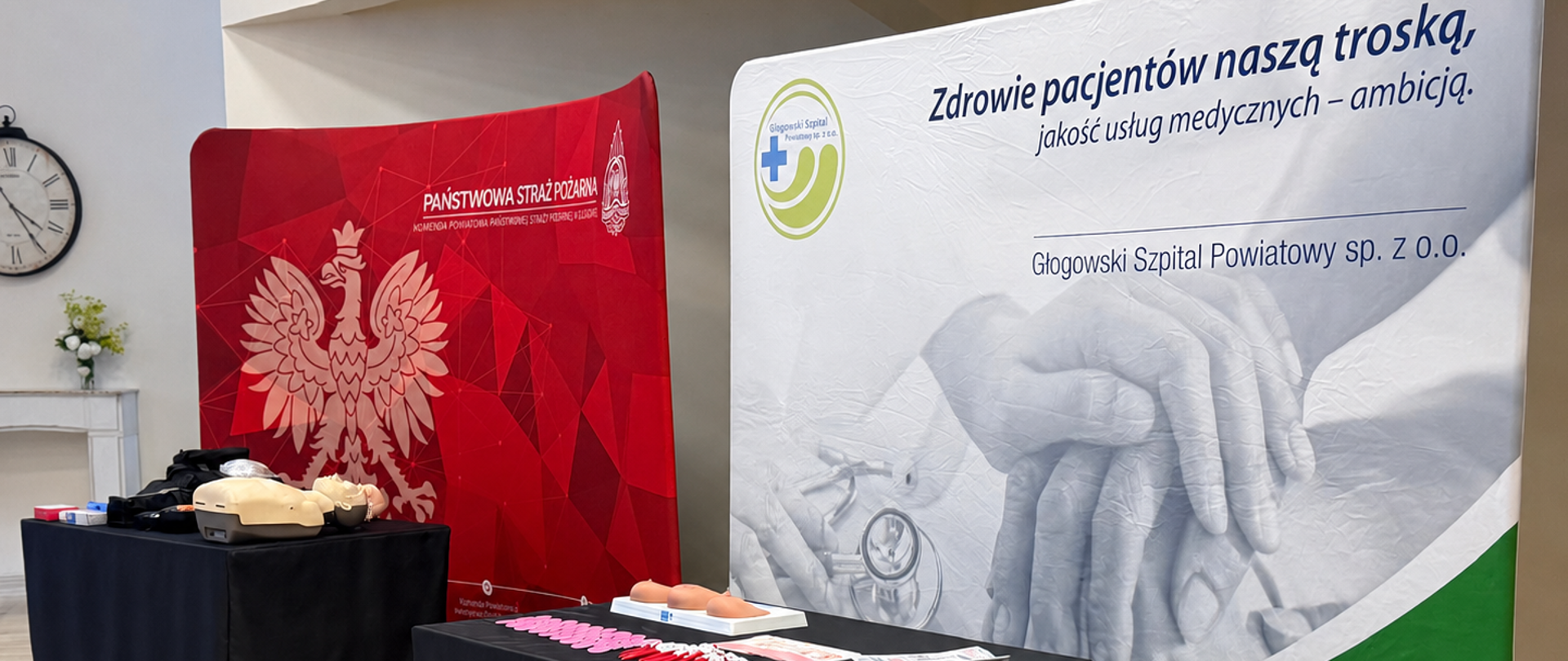 Stanowisko informacyjno-edukacyjne podczas wydarzenia „Bezpieczna MAMA”, które odbyło się 25 maja 2026 roku w Miejskim Centrum Integracji „Jubilat” w Głogowie. Na zdjęciu widoczne są dwa roll-upy promocyjne: Państwowej Straży Pożarnej oraz Głogowskiego Szpitala Powiatowego. Przed nimi ustawiono stoły z materiałami edukacyjnymi i fantomami do nauki pierwszej pomocy. Wydarzenie poświęcone było bezpieczeństwu oraz zdrowiu kobiet, ze szczególnym uwzględnieniem profilaktyki zdrowotnej i edukacji w zakresie ratownictwa.