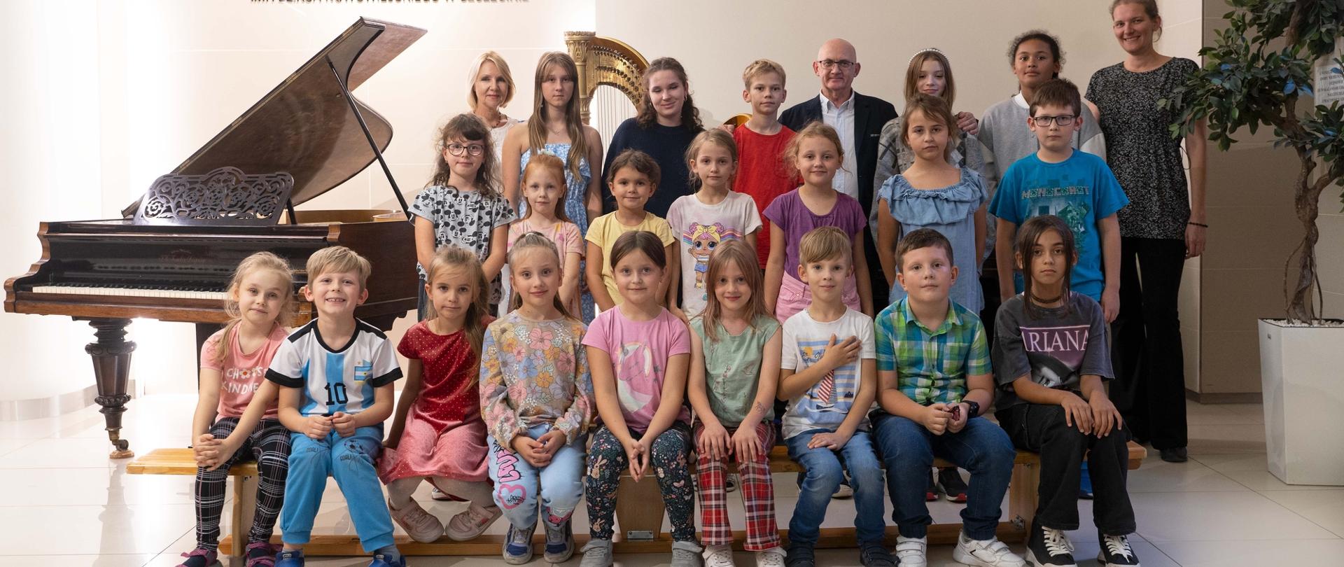 zdjęcie przedstawia nowych uczniów szkoły pierwszego stopnia we wspólnym ujęciu razem z dyrekcją w holu szkoły, w tle fortepian i logo szkoły na ścianie