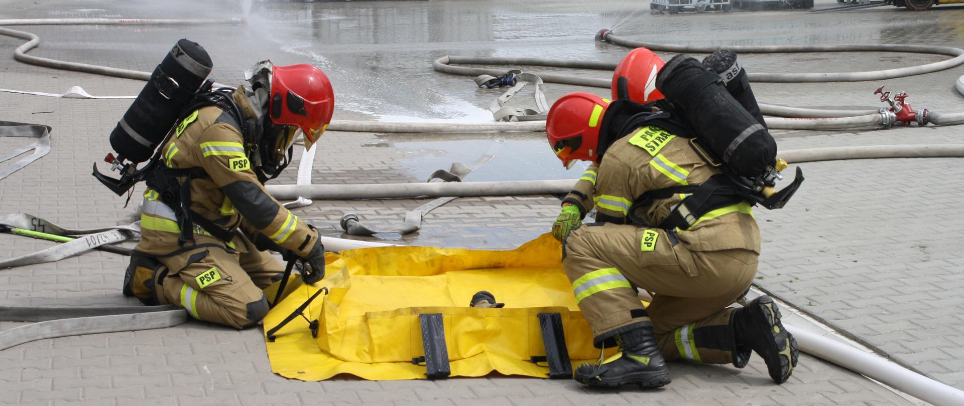 Strażacy PSP w pełnym ubraniu bojowym składają leżącą na placu firmy żółtą wannę wychwytową . W tle kurtyny wodne, wózek widłowy oraz mauzery.