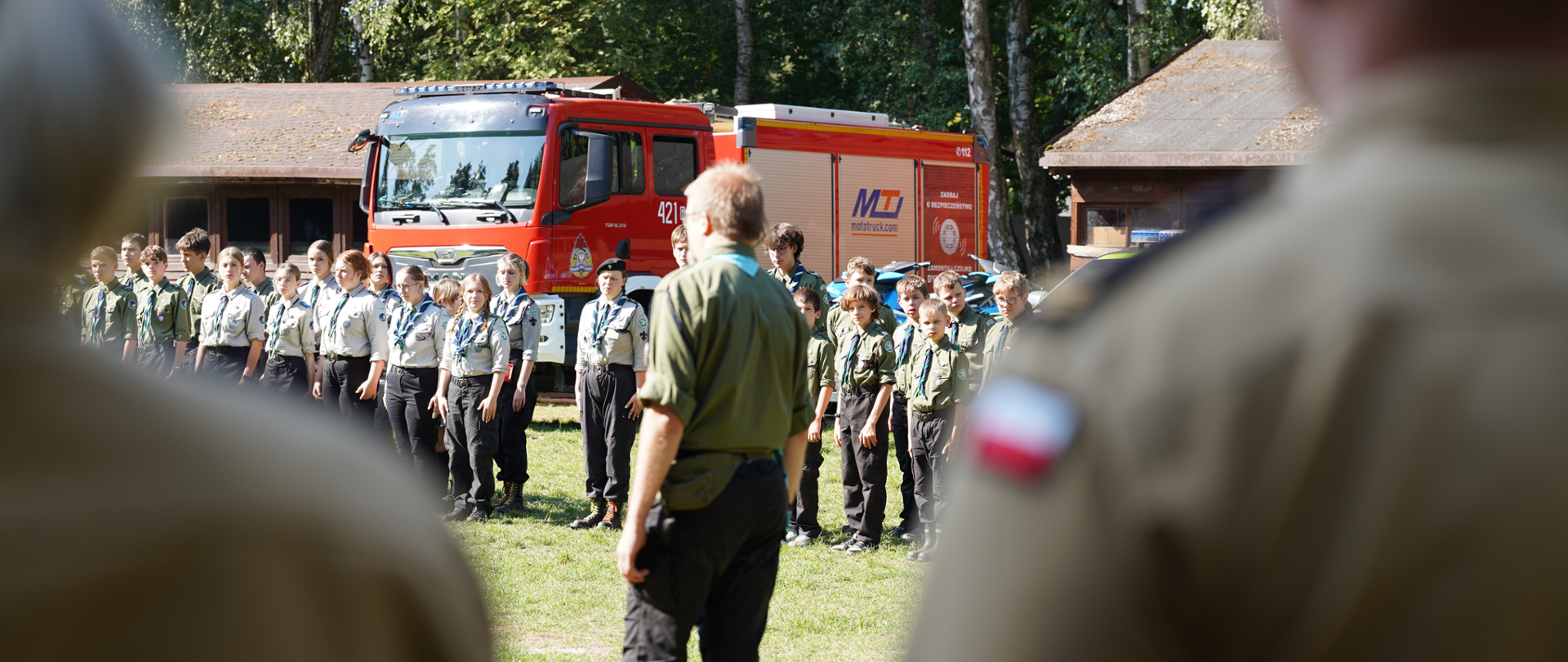 Zdjęcie przedstawia grupę młodych harcerzy stojących na trawie przed czerwonym wozem strażackim. W centrum kadru, tyłem do aparatu, stoi dorosły mężczyzna prowadzący zbiórkę. Na pierwszym planie, częściowo rozmyte, widoczne są sylwetki dwóch osób w mundurach. W tle znajdują się drewniane budynki i las.
