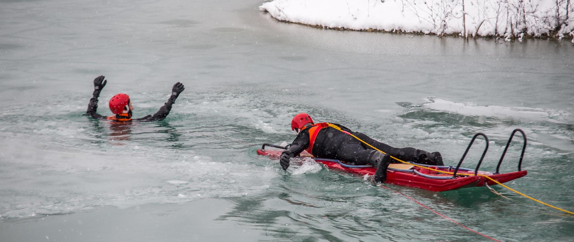 Zdjęcie przedstawia dwóch strażaków w ubraniach wypornościowych (piankach) w pełnej asekuracji podczas ćwiczeń lodowych. Jeden ze strażaków to osoba tonąca (pośród lody na akwenie) a drugi podpływa na desko-saniach lodowych podpływa do osoby poszkodowanej.