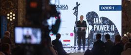 W zaciemnionej sali pod ekranem z napisem Dzień 1 - Jan Paweł II stoi minister Czarnek i mówi do mikrofonu na stojaku.