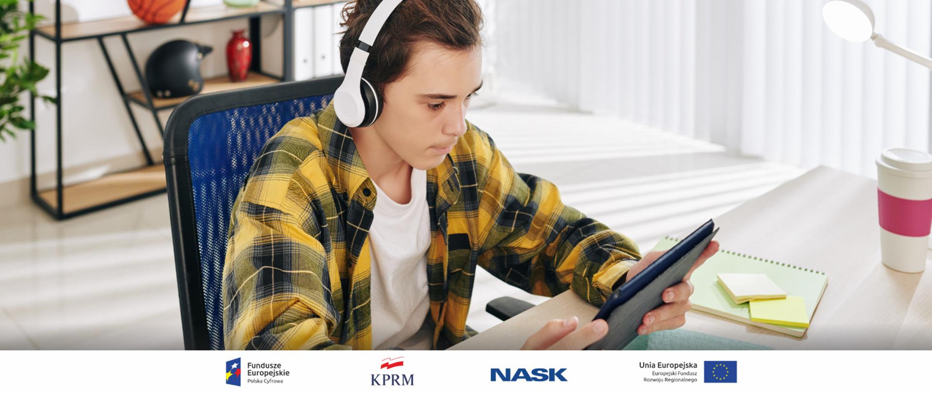 Zdjęcie nastolatka w słuchawkach siedzącego przy biurku - w rękach trzyma tablet, na którym coś ogląda.