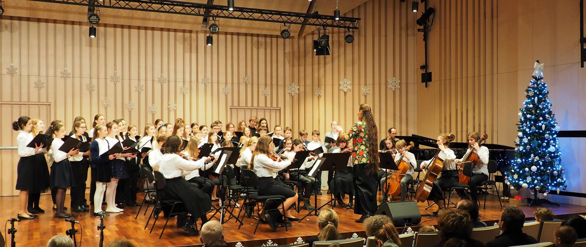 Scena w kolorze berzowym-brązowym, na której występuje orkiestra smyczkowa i chór szkolny pod dyrekcją Agaty Orłowskiej-Półtorak po prawej stronie choinka świąteczna