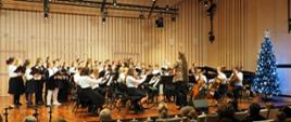 Scena w kolorze berzowym-brązowym, na której występuje orkiestra smyczkowa i chór szkolny pod dyrekcją Agaty Orłowskiej-Półtorak po prawej stronie choinka świąteczna