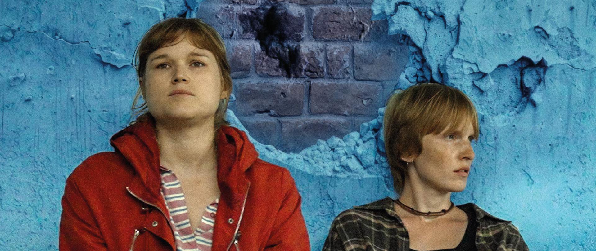 El cartel de la película “Dos hermanas” muestra a dos mujeres jóvenes sentadas en el suelo, frente a una pared azul descascarada con una gran parte del yeso desprendido, a través de la cual se ven los ladrillos. La mujer de la izquierda lleva una chamarra roja y mira hacia el frente con una expresión seria y pensativa, mientras que la mujer de la derecha tiene el cabello corto y claro, viste una camisa oscura a cuadros y mira hacia un lado. Sobre ellas, en grandes letras amarillas, aparece el título “DOS HERMANAS” y más arriba la información de que es una película dirigida por Łukasz Karwowski. El cartel anuncia la proyección el 26 de febrero a las 18:00 horas en la Alianza Francesa Polanco, en México, y que la entrada es gratuita. En la parte inferior se encuentran los logotipos de las instituciones, entre ellas las embajadas de Polonia y Ucrania, así como la Unión Europea.