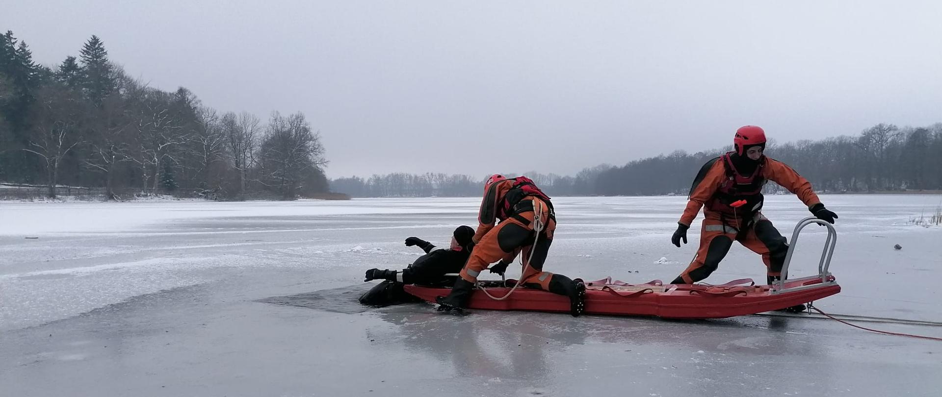 Zdjęcie przedstawia zamarznięte jezioro z widocznym podłużnym przeręblem w którym znajduje się dwóch strażaków ubranych w żółte skafandry wypornościowe. Jeden ze strażaków znajduję się w wodzie zaś dwóchi na czerwonych saniach wodno-lodowych z przypiętą liną. W tle widać las.
