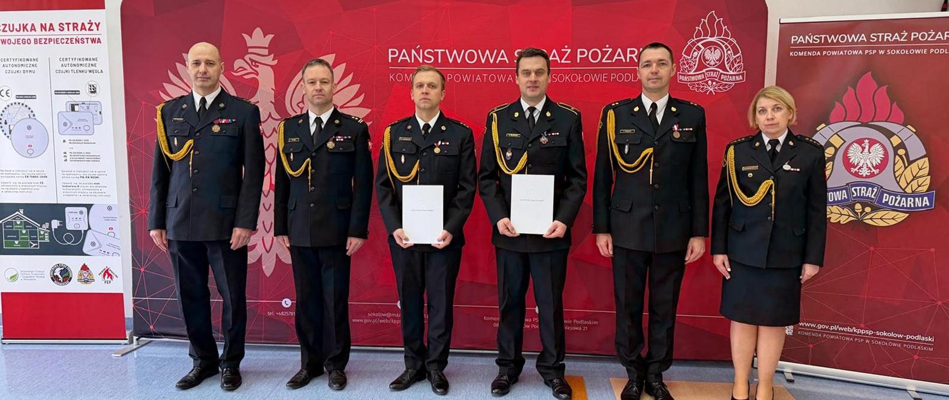 Zdjęcie przedstawia funkcjonariuszy KP PSP Sokołów Podlaski w umundurowaniu galowym stojących w jednym rzędzie. Stoją na tle czerwonych bilbordów o tematyce strażackiej. Nowo mianowani dowódcy trzymają przed sobą akty mianowania w kolorze białym.