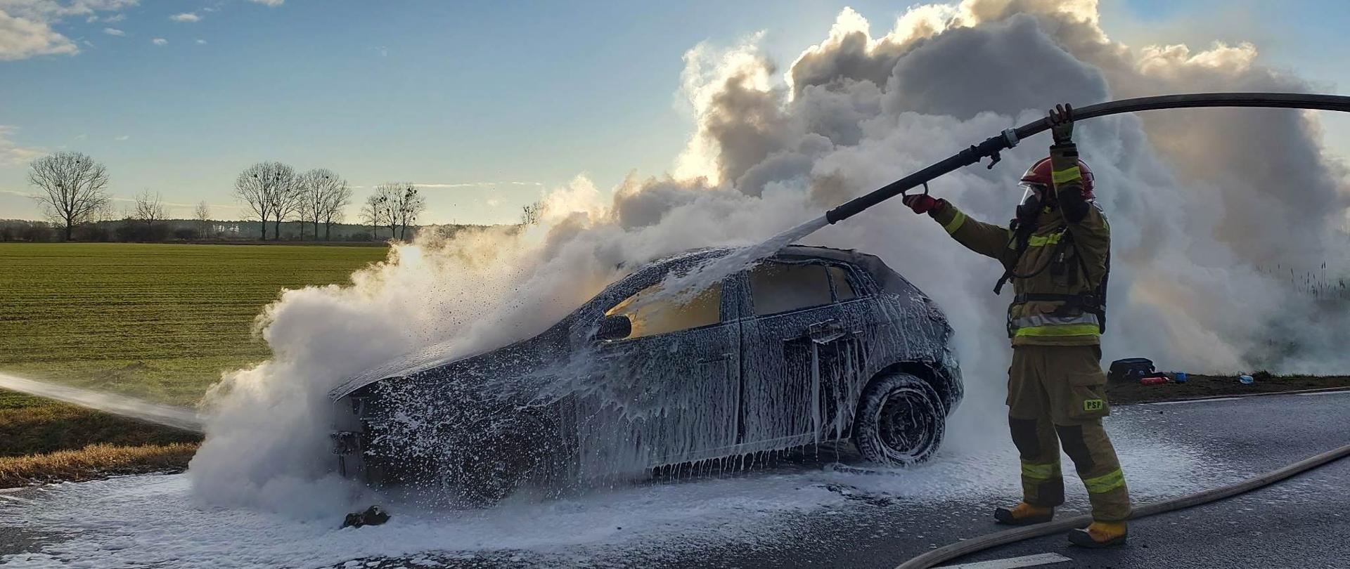 Pożar samochodu. Droga wojewódzka nr 434 w okolicach miejscowości Roszkówko. Na tle pola i drzew pali się ciemny samochód osobowy stojący na jezdni. Obok samochodu stoi strażak, który trzymając nad głową wąż i prądownicę, przy użyciu piany gasi pożar. Z pojazdu wydobywa się gęsty biały dym.