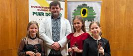trzy dziewczynki ze skrzypcami i jeden mężczyzna, za nimi dwa banery i drewniana ściana