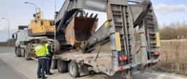 Ciągnik siodłowy z podpiętą naczepą stoi w punkcie kontrolnym. Obok naczepy przewożącej dużą koparkę gąsienicową stoi kierowca i umundurowany inspektor warmińsko-mazurskiej Inspekcji Transportu Drogowego. Funkcjonariusz mierzy wysokość naczepy z ładunkiem za pomocą specjalnego narzędzia.