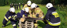 Widok z przodu. Drużyna strażaków OSP przecina leżący na podkładzie z pięciu europalet element drewniany przy pomocy ręcznej piły poprzecznej do cięcia drewna. W tle kolejna drużyna oczekująca na start. 