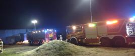 Na zdjęciu widać pojazdy straży pożarnej w kolorze czerwonym. Zdjęcie w porze nocnej, na terenie działań ratowniczo-gaśniczych. W tle zabudowania. Rozwinięte linie gaśnicze od pojazdów.