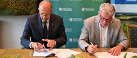 Na zdjęciu na tle banera z logo Narodowego Funduszu Ochrony Środowiska i Gospodarki Wodnej siedzą przy stole od lewej Zastępca Prezesa Narodowego Funduszu Ochrony Środowiska i Gospodarki Wodnej Robert Gajda i Prezes Zarządu Przedsiębiorstwa Wodociągów i Kanalizacji Janusz Głuszek. Zdjęcie przedstawia moment podpisania umowy. 