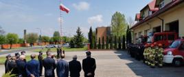 Zdjęcie przedstawia plac przed budynkiem komendy, wyłożony kostką. Po prawej stronie stoją w dwuszeregu strażacy w galowym oraz bojowym umundurowaniu, a za nimi stoją samochody strażackie. Po lewo znajdują się zaproszeni goście, mównica, przy której stoją dwie osoby, mężczyzna i kobieta w galowym umundurowaniu, a następnie widać masz z flagą i stojący przy niej trzej strażacy w galowym umundurowaniu.