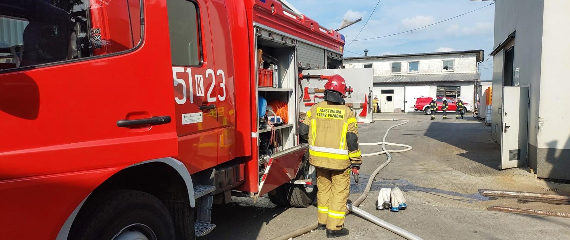 Zdjęcie przedstawia strażaka oraz samochód pożarniczy biorący udział w działaniach ratowniczo-gaśniczych. W centralnej części zdjęcia można zauważyć rozwinięte linie wężowe. 
