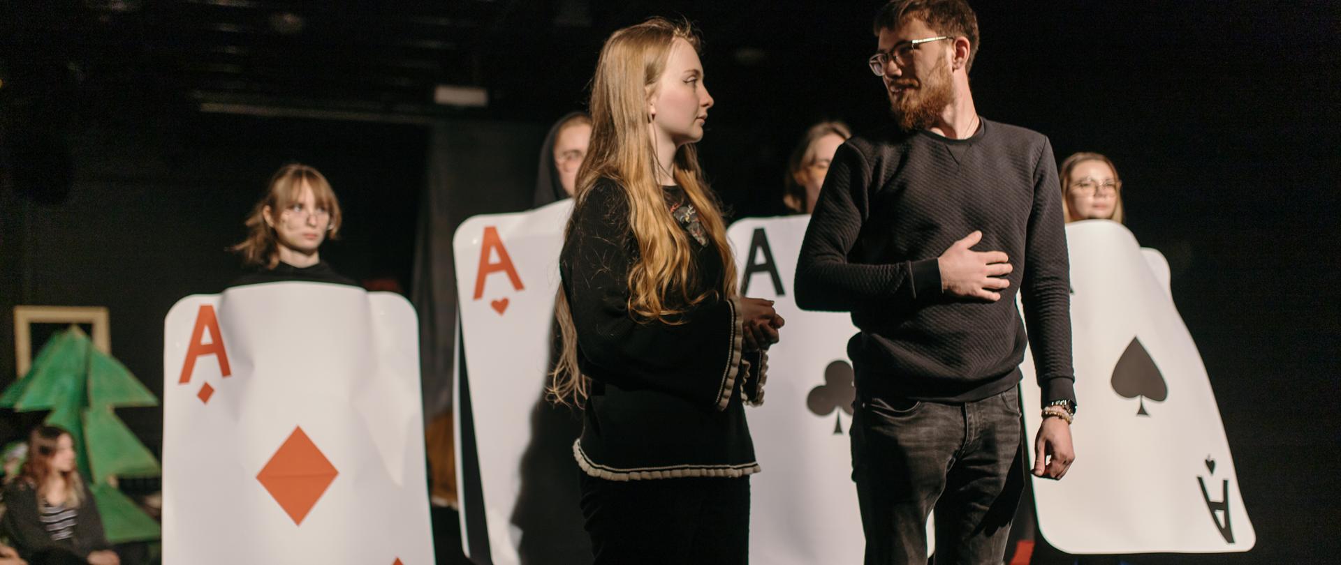 Mężczyzna i kobieta na pierwszym planie, w tle słabo widoczne postaci w strojach karcianych.