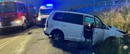 Pora nocna samochód osobowy koloru białego z uszkodzonym przodem i tyłem przez otwarte prawe przednie drzwi widać zdetonowaną kurtynę boczną oraz poduszki powietrzne pasażera i kierowcy. Za samochodem stoi biała karetka pogotowia z łączonymi światłami mijania i niebieską sygnalizacją świetlną z prawej strony karetki zaparkowany czerwony samochód Straży Pożarnej z włączonymi światłami mijania oraz niebieską sygnalizacją świetlną oraz wysuniętym masztem oświetlającym teren działań ratowniczych. Pomiędzy samochodami Straży Pożarnej i pogotowia strażak w ubraniu specjalnym niosący na plecach torbę medyczną koloru czerwonego. Na skarpie za barierkami stoi osoba przyglądająca się zdarzeniu.