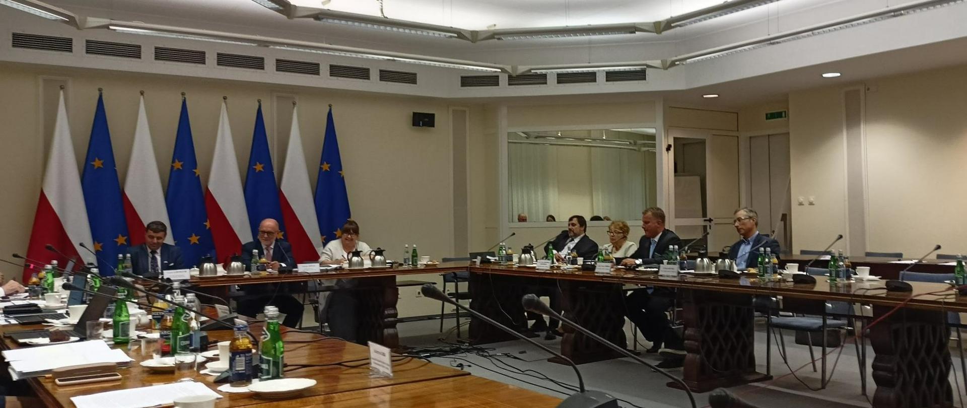 Na zdjęciu przy długich stołach w sali konferencyjnej siedzi około dziesięciu osób. Przed nimi na stołach leżą kartki, napoje w butelkach, talerzyki i filiżanki oraz mikrofony. Wszyscy są elegancko ubrani, panowie w garniturach. Za nimi stoją flagi Polski i Unii Europejskiej. 