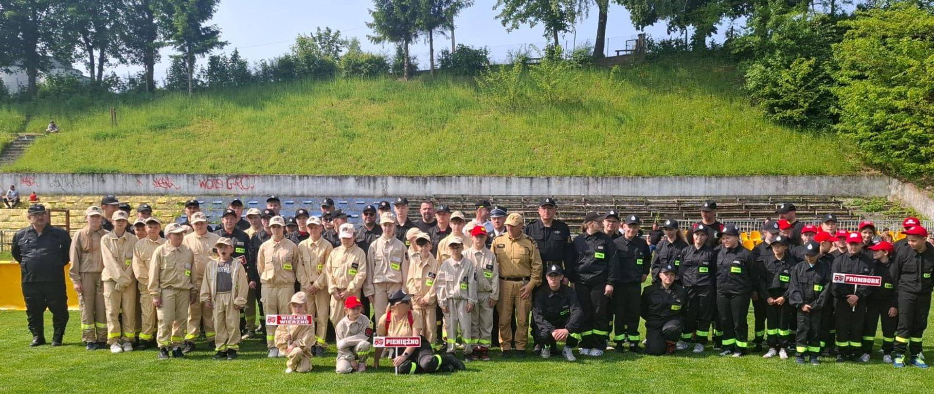 Na zdjęciu widzimy dużą grupę młodych strażaków i opiekunów, stojących wspólnie na zielonym boisku, podczas wydarzenia związanego z zawodami Młodzieżowych Drużyn Pożarniczych (MDP). Uczestnicy są podzieleni na grupy w różnych mundurach: jasno-beżowych i czarnych z żółtymi pasami odblaskowymi. Na pierwszym planie widać dzieci trzymające tabliczki z nazwami drużyn: „Wielkie Wierzno”, „Pieniężno” i „Frombork”, co sugeruje, że to lokalne zawody gminne lub powiatowe. Uczestnicy mają czapki strażackie, niektórzy również bluzy z napisem „STRAŻ”. W tle – betonowe trybuny, zarośnięte skarpy i graffiti na murze. Pogoda jest słoneczna, a atmosfera wygląda na radosną i zorganizowaną.