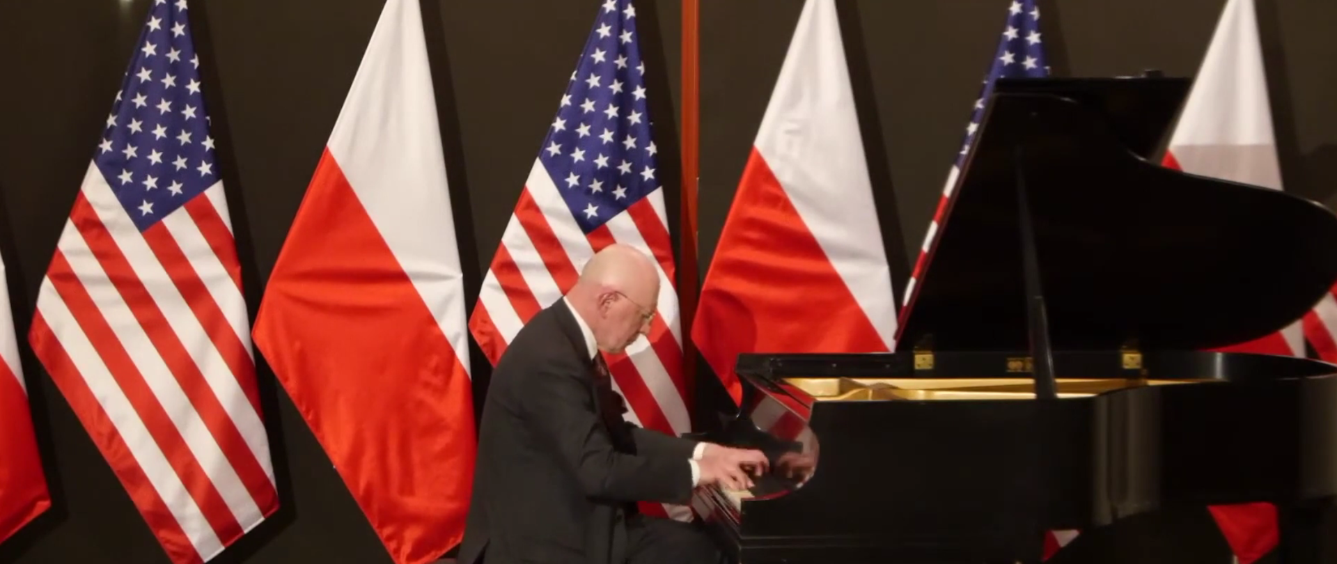 Marek Żebrowski gra na fortepianie utwór Ignacego Paderewskiego, Cracovienne Fantastique, Op. 14 NO. 6. Zdjęcie w tle przedstawia flagi polską i amerykańską. .