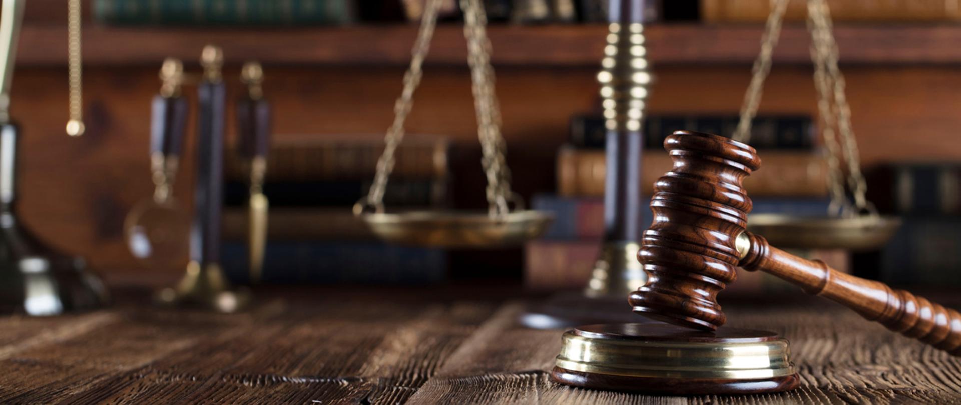 Blat biurka. Na nim drewniany młotek sędziowski na podstawce, w tle ozdobna waga i książki.