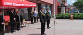 Na pierwszym planie widzimy dwóch mężczyzn, strażaków stojących bokiem do kadru. Ubrani w ciemne mundury galowe, czapki na głowach, na mundurach widać także sznury koloru złotego. Mężczyzna z lewej stoi przed mikrofonem i przemawia. W tle widzimy trzech strażaków podobnie ubranych stojących w rzędzie, trzymają sztandar. Z lewej strony kadru widzimy namiot koloru czerwonego z białym napisem: komenda miejska państwowej straży pożarnej w Kaliszu. Pod namiotem widzimy trzech mężczyzn, cywili ubranych w czarne garnitury, białe koszule, ciemne krawaty. Dalej w tle widzimy budynek straży pożarnej koloru łososiowego, przeszklone wejście główne.