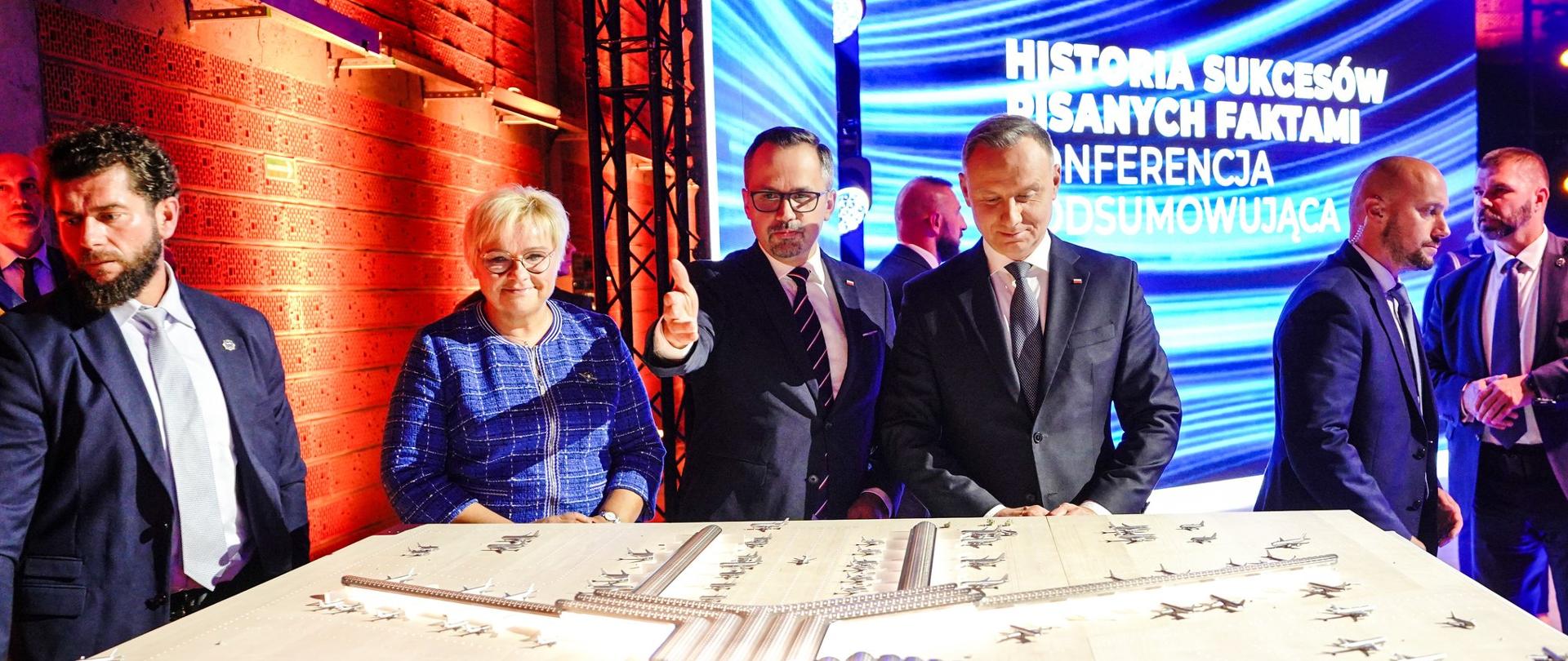Prezydent RP, Andrzej Duda, wiceminister funduszy i polityki regionalnej, Marcin Horała i uczestnicy konferencji na konferencji podsumowującej prace przy CPK, przyglądają się makiecie portu