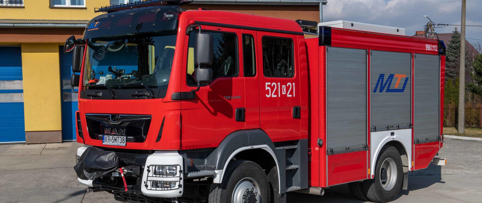 Dwuosiowy średni samochód ratowniczo gaśniczy marki MAN koloru czerwonego z białym zderzakiem z wyciągarką z przodu, na tle bram wjazdowych do garażu od strony placu wewnętrznego.