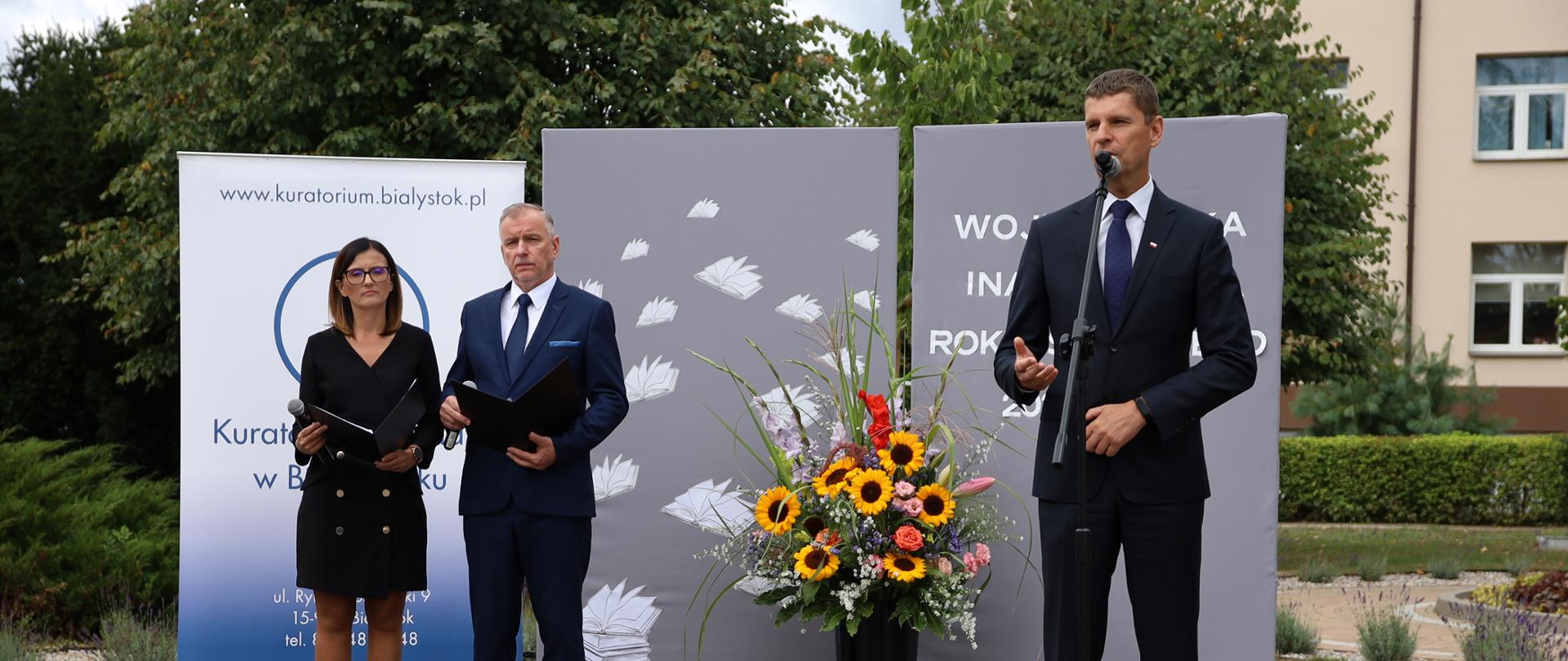 Minister Dariusz Piontkowski przemawia na scenie za nim kobieta i mężczyzna trzymający w dłoniach otwarte dokumenty