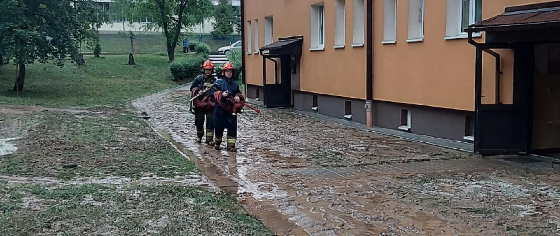Zdjęcie przedstawia po prawej stronie część bloku czteropiętrowego z dwoma wejściami na klatki schodowe. Przed budynkiem na chodniku płynie woda i osadza się duże ilości błota. Po chodniku idzie dwóch strażaków ubranych w mundury specjalne i czerwone hełmy w rękach trzymają węzę pożarnicze oraz klucze do hydrantu.Za blokiem widać dwa drzewa i schody.