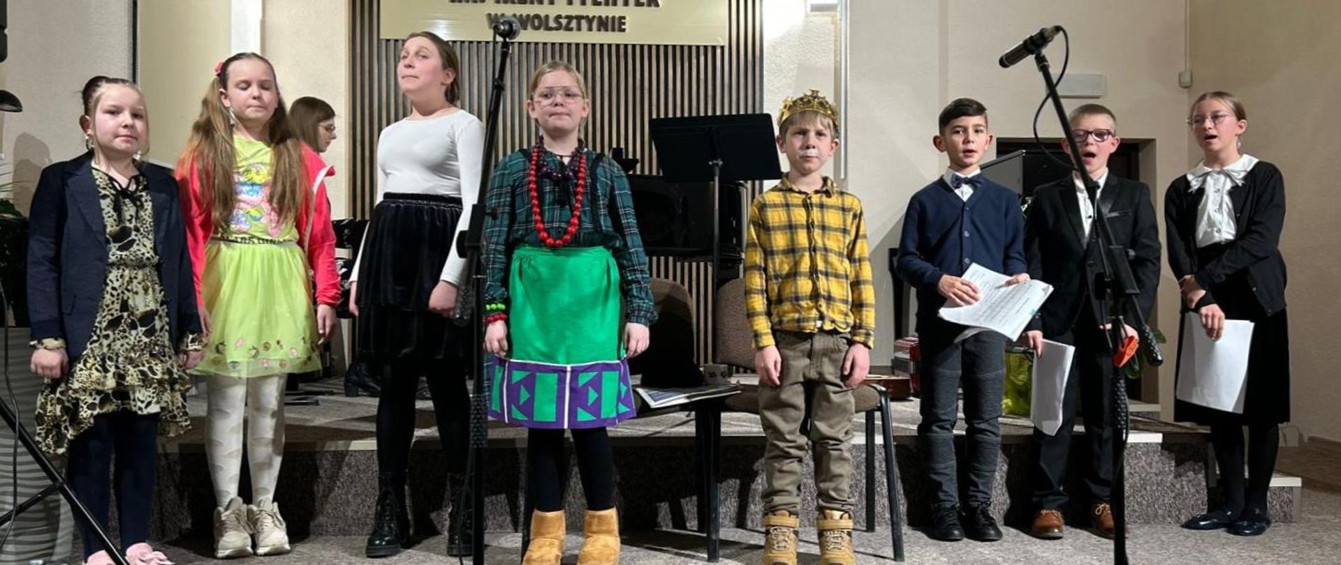 Dzieci z młodszej grupy chóralne stoją na scenie i śpiewają piosenkę.