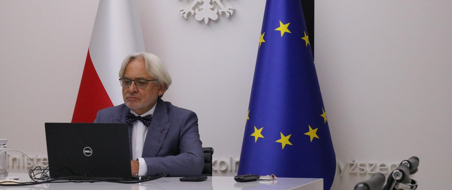 Minister W. Maksymowicz siedzi przed monitorem przy biurku, w tle flagi polska i UE.