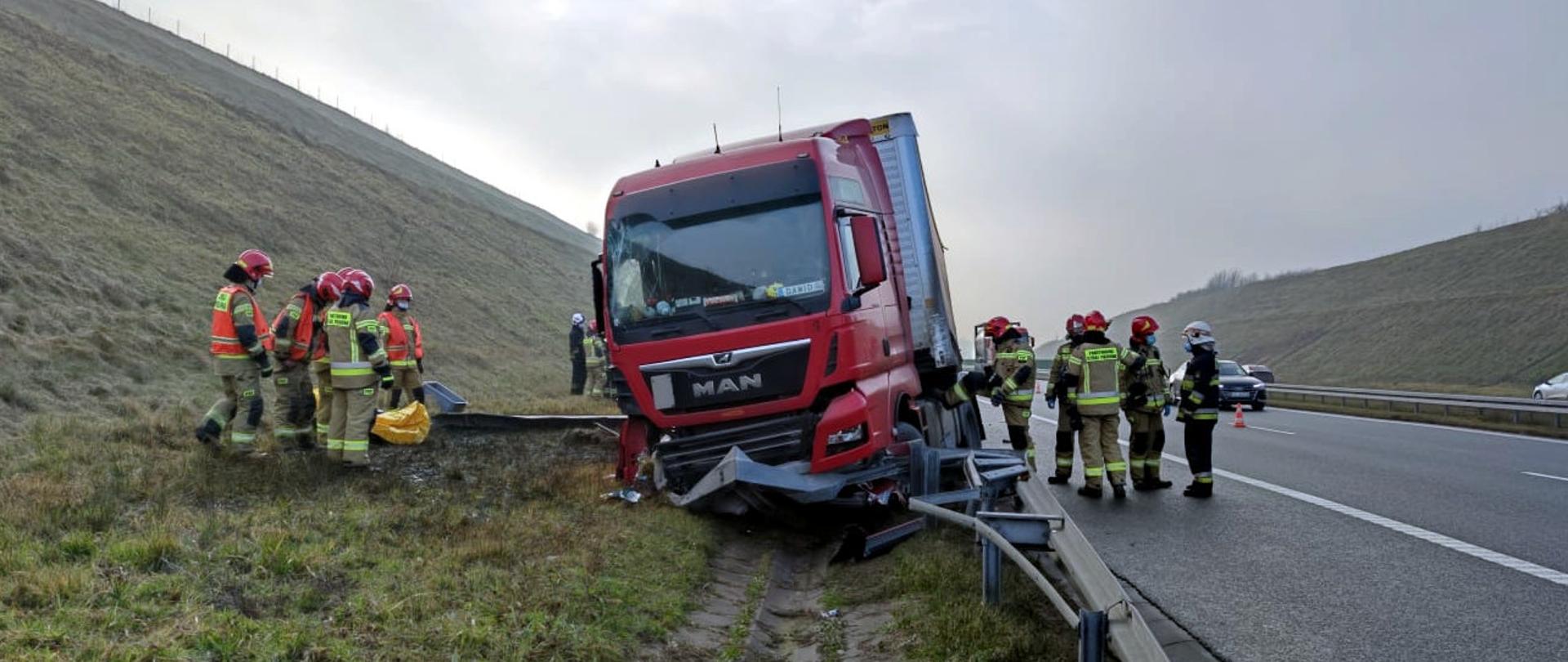 Zdjęcie przedstawia samochód ciężarowy, który po staranowaniu barierki utknął w rowie. Wokół strażacy realizujący czynności ratownicze. Z lewej strony widać zarośnięty wysoki nasyp, a z prawej autostradę A1. 