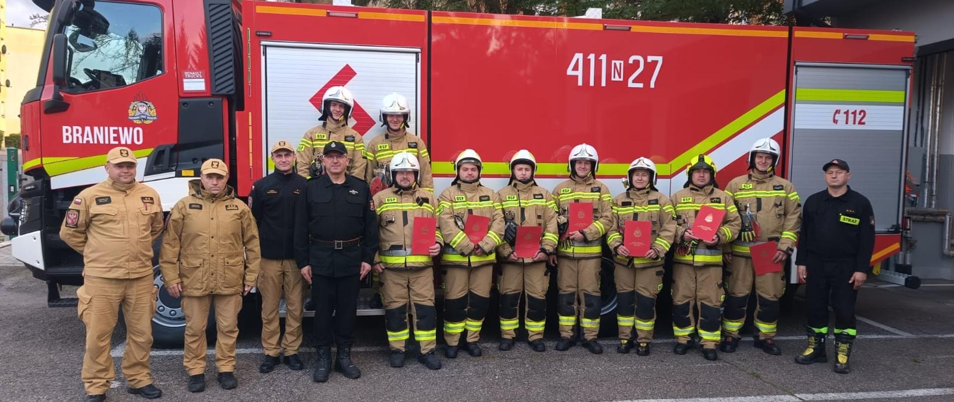 Na tle dużego czerwonego samochodu strażackiego stroją w jednym szeregu strażacy w mundurach i hełmach oraz strażacy PSP