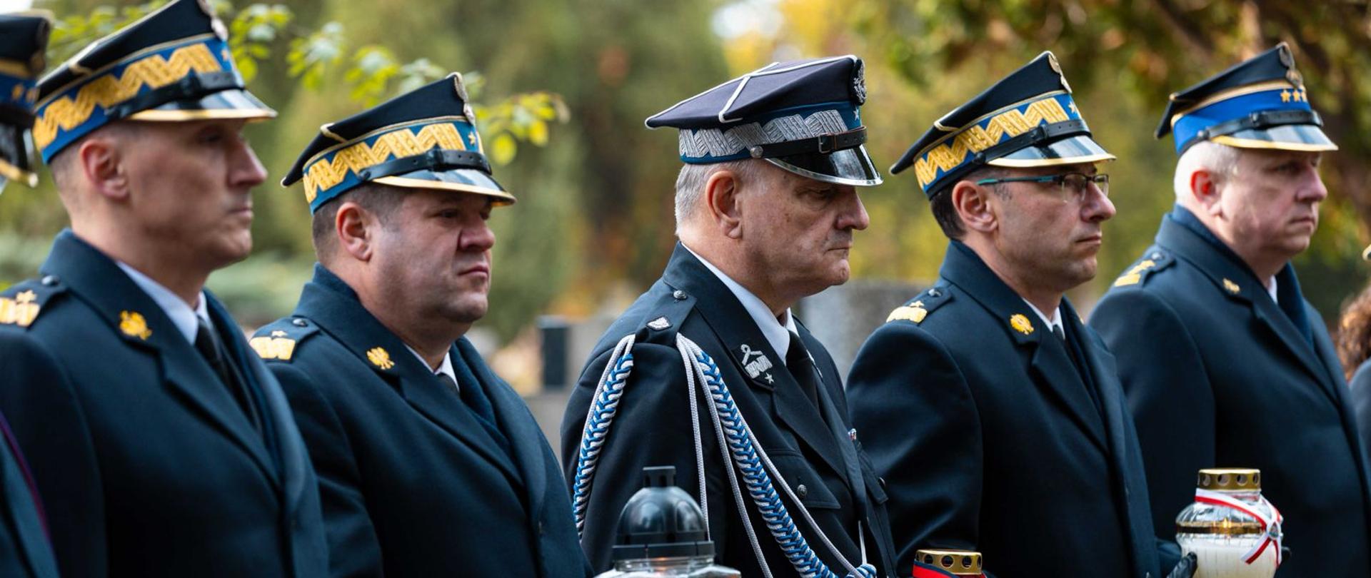 Oficerowie PSP w galowych mundurach stoją w szeregu, trzymając zapalone znicze w barwach narodowych podczas ceremonii pamięci.