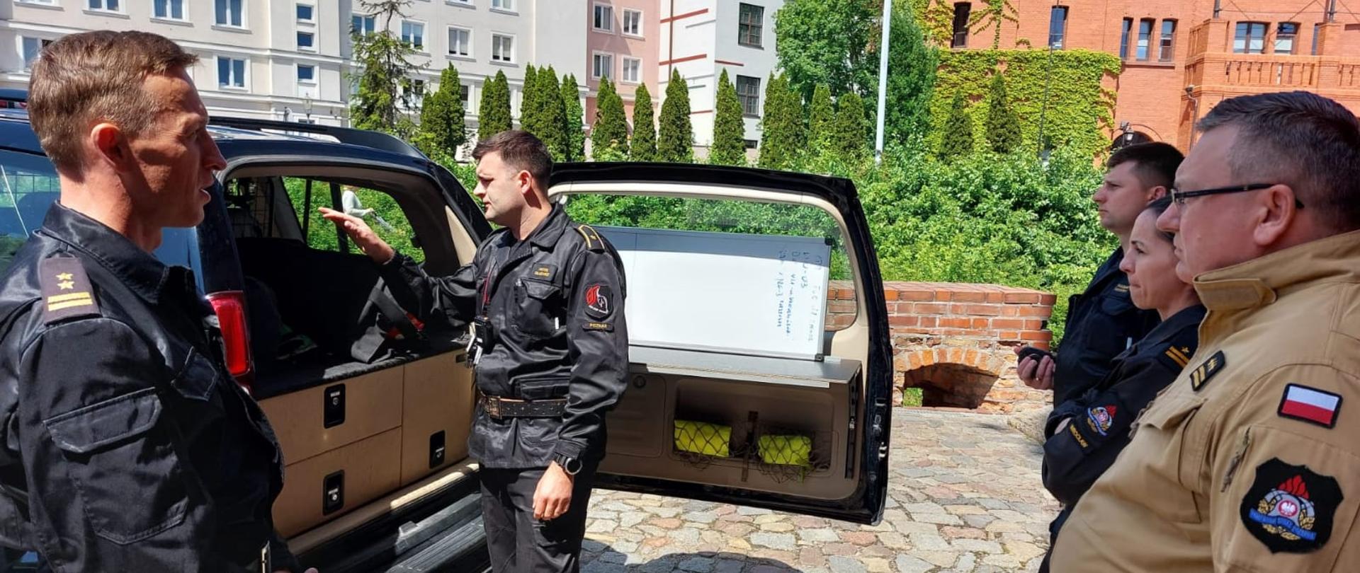 strażacy oglądają samochós strażacki w tle zielone krzewy i budynki mieszkalne