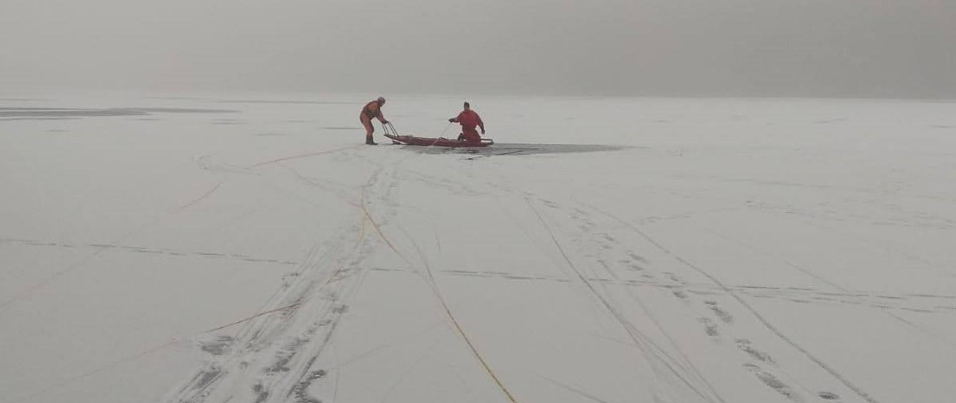 Zamarznięta tafla jeziora, pokryta śniegiem, w tle zdjęcia dwóch strażaków ubranych w kombinezon do pracy w wodzie, przy wykutym przeręblu zabezpieczonych saniami lodowymi i linką. 