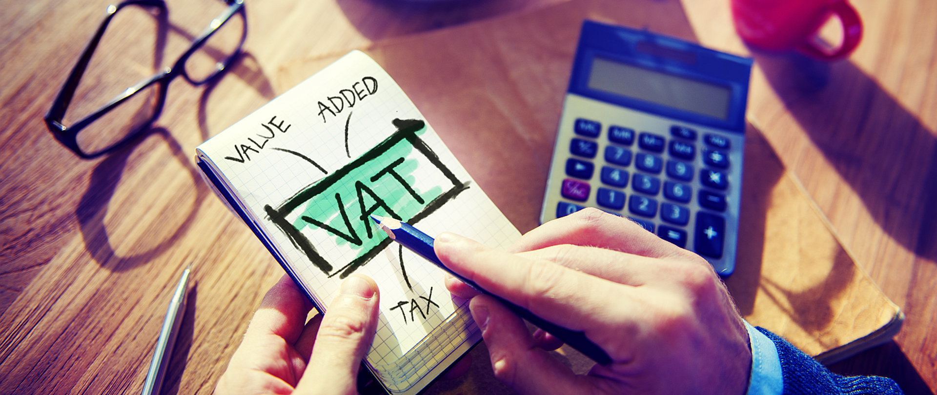 Notes na którym widnieje napis VAT w tle widać blat stołu na którym jest kalkulator okulary i długopis