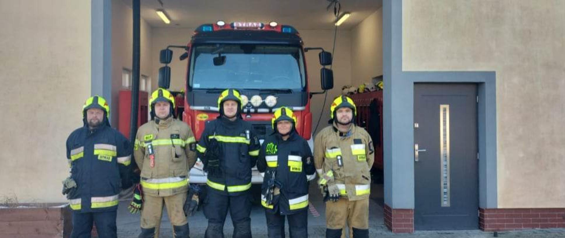 Na zdjęciu widzimy strażaków poddanych Inspekcji Gotowości Operacyjnej na tle remizy i samochodu pożarniczego.
