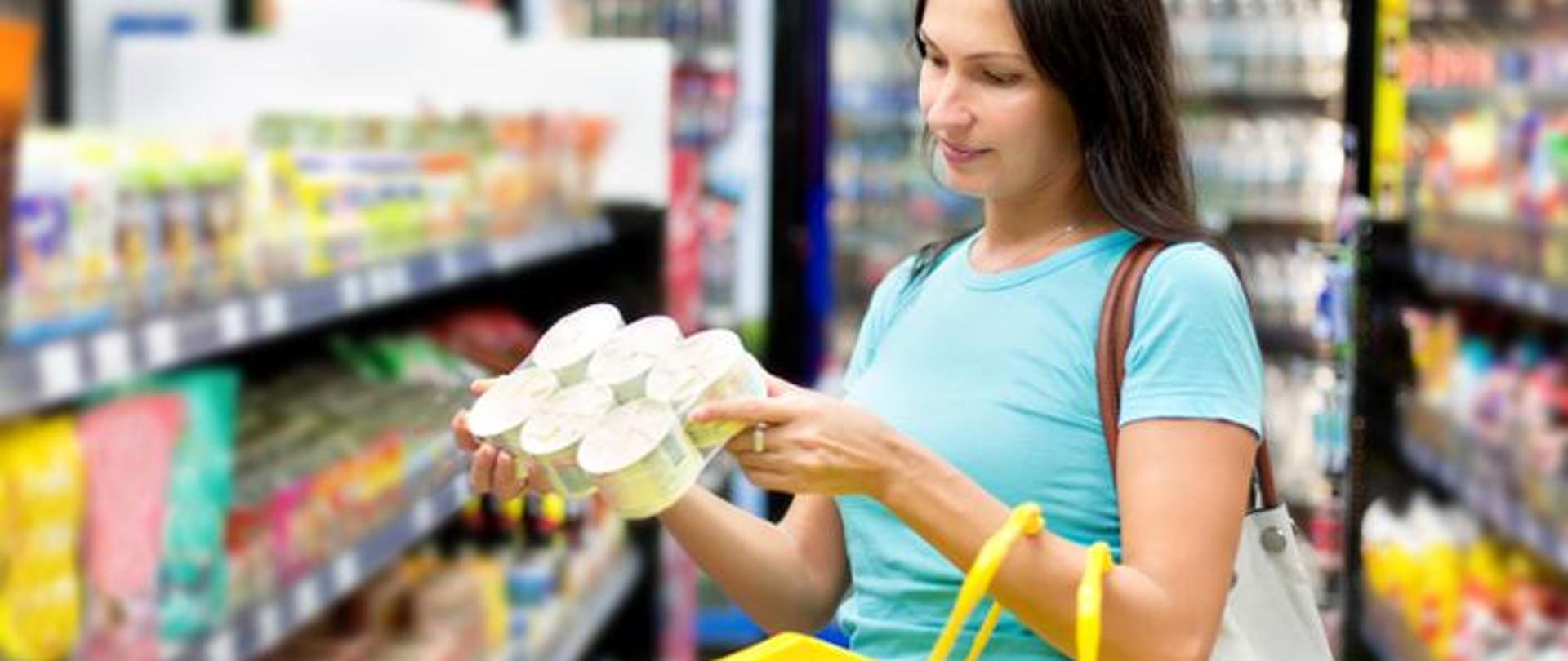 Kobieta ciemne długie włosy niebieska koszulka trzyma w ręku produkty spożywcze czyta etykiety na produkcie. w tle regały sklepowe