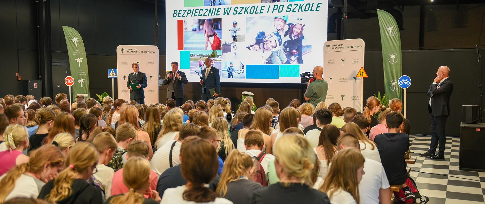 Widok z tyłu na wypełnioną kolorowo ubranymi dziećmi salę, przed nimi wielki ekran z napisem Bezpiecznie w szkole i po szkole.