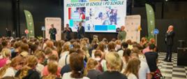 Widok z tyłu na wypełnioną kolorowo ubranymi dziećmi salę, przed nimi wielki ekran z napisem Bezpiecznie w szkole i po szkole.