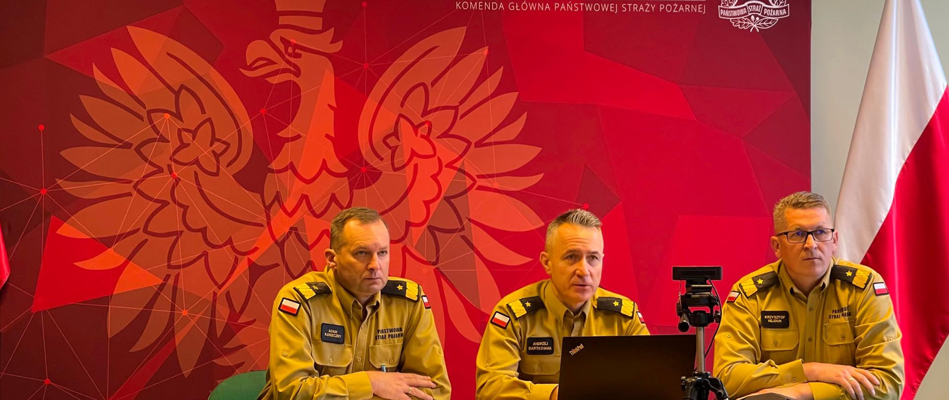 siedzą przy stole podczas wideokonferencji Komendant Główny PSP (po środku) po lewo i prawo siedzą jego zastępcy, w tle na ściance koloru czerwonego widoczny symbol orła i napis PAŃSTWOWA STRAŻ POŻARNA , KOMENDA GŁÓWNA PSP, przed nimi laptop