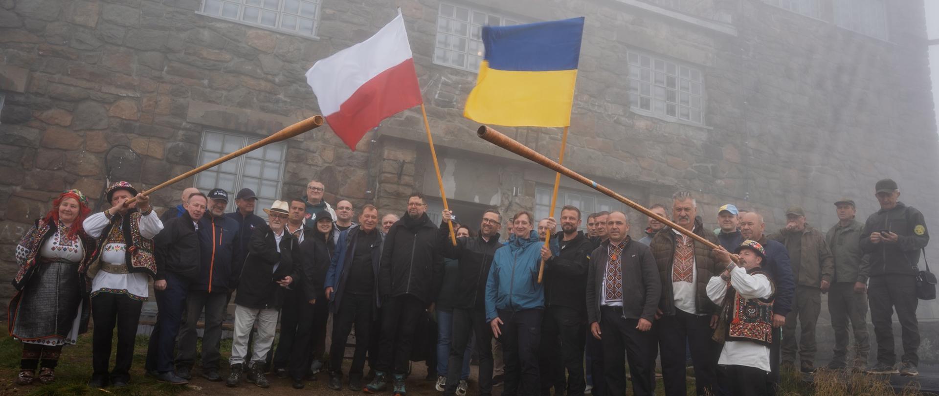 Mglisty dzień, pod budynkiem z kamiennymi ścianami stoi grupa kilkudziesięciu osób, po bokach dwaj mężczyźni w ludowych kolorowych strojach grają na długich trąbach, pośrodku grupy minister Kulasek trzyma polską flagę, obok niego mężczyzna w niebieskiej kurtce trzyma flagę Ukrainy.